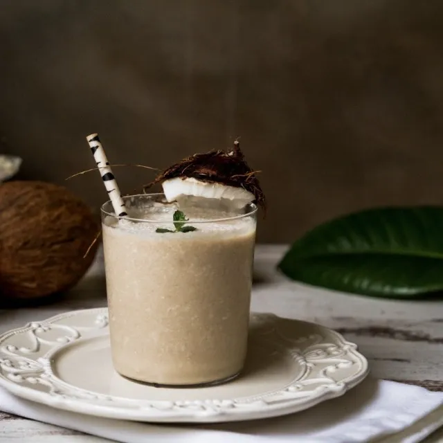 Milky liquid in glass with straw on a table decorated with coconunts and leaves 