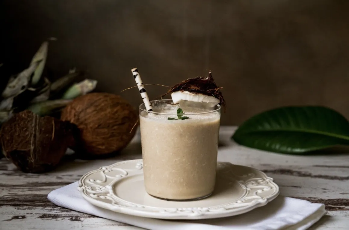 Milky liquid in glass with straw on a table decorated with coconuts and leaves 