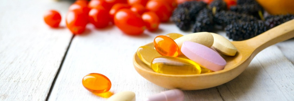 Assorted pills and capsules on a wooden spoon with fresh fruits in the background, illustrating health supplements.